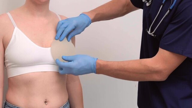 Plastic surgeon examining a female patient and holding breast implants near her body in a clinic, healthcare advertising, plastic surgery, patient examination.