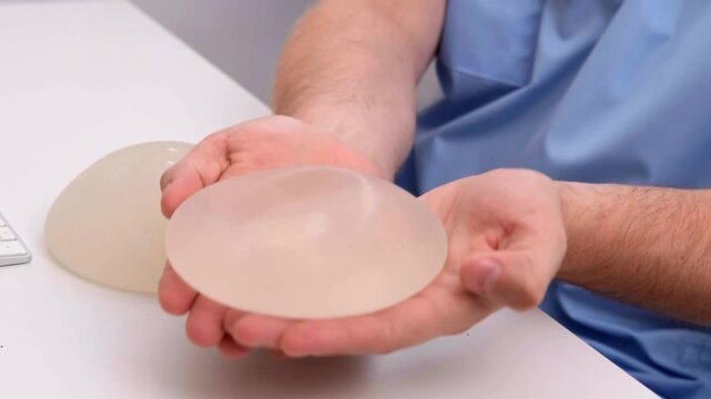Plastic surgeon demonstrating breast implants by holding them in gloved hands at a medical clinic, healthcare advertising, plastic surgery, aesthetic medicine.