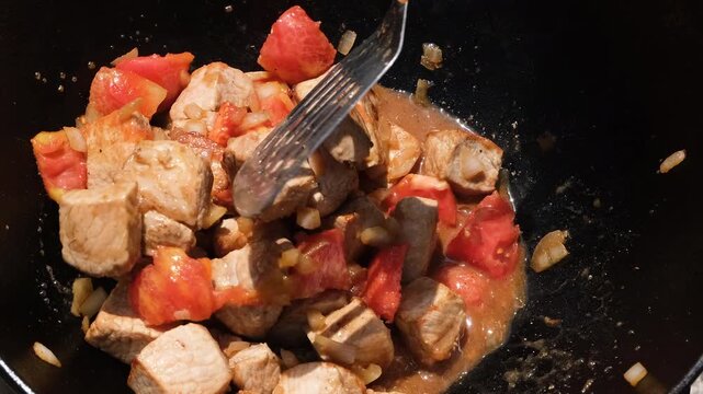 Cooking pork meat with fresh tomatoes and onions in a traditional cauldron over an open fire outdoors.