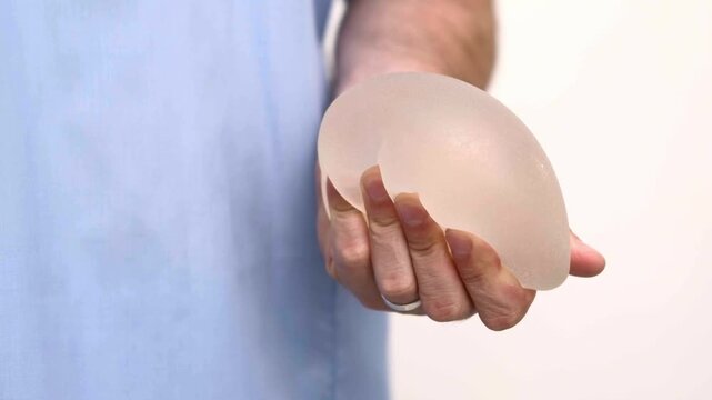 Surgeon holding a single anatomical breast implant in scrubs, plastic surgery clinic consultation, aesthetic medicine and augmentation mammoplasty patient education.