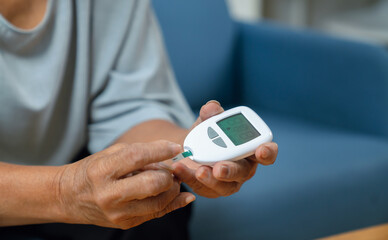 Elderly Asian woman using a blood glucose meter while sitting on a sofa at home, diabetes care and health monitoring. © sitthiphong