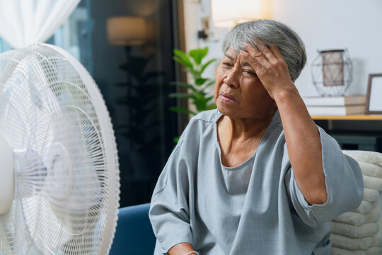 Elderly Asian woman sitting on a sofa, feeling stressed from the heat while turning on a fan to cool down in an overheated living room.