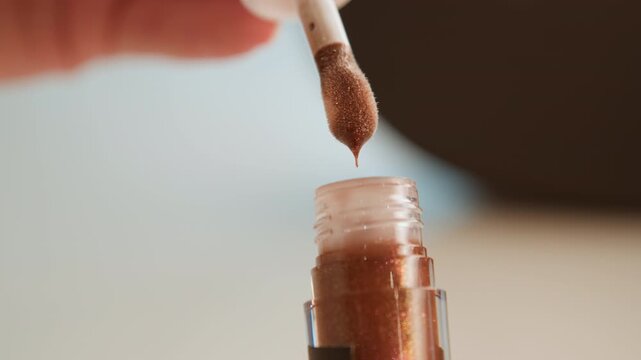 Close-up motion of doe-foot wand rising from clear bottle filled with shimmery brown-gold cosmetic liquid, with viscous product stretching from tip, glossy residue on wall, soft studio light