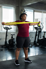 Fototapeta premium Plus size African American woman stretching arms with green resistance band in modern gym, fitness lifestyle photo for shoulder mobility, healthy workout, body confidence and wellness motivation.