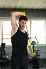 Fototapeta premium Adult Caucasian man holding green resistance band overhead in modern gym, shoulder mobility stretch before workout, active male athlete wellness routine for personal training and gym membership ads