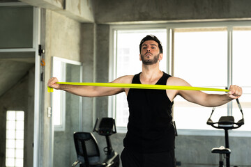 Fototapeta premium Adult Caucasian man extending green resistance band across upper body in modern gym, shoulder mobility stretch before workout, active male athlete wellness routine for personal training gym ads