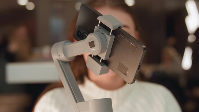 Close-up view shows smartphone secured on gimbal stabilizer with visible arm, joint, and subtle rotational movement while woman remains softly blurred behind it in warm indoor light, suggesting