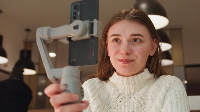 Young woman uses smartphone gimbal inside cozy caf , adjusting handheld stabilizer while speaking, smiling, and creating engaging social in warm natural light with relaxed indoor atmosphere