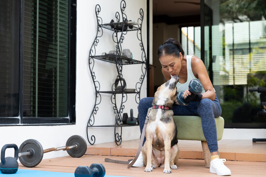 A healthy senior woman shares an affectionate moment with her pet dog while holding a dumbbell during a home workout. The image represents healthy aging, emotional connection, and an active lifestyle