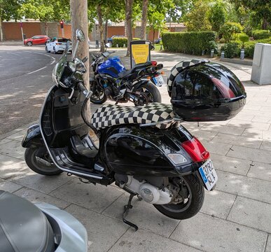 Vista lateral de moto scooter estilo retro Vespa GTS Super con ba&uacute;l trasero y Checkers Seat Cover, estacionada en Madrid, Espa&ntilde;a, 5 de junio de 2026