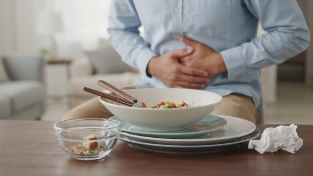Person holding their stomach in pain while sitting at a table with a stack of dirty dishes and a crumpled napkin after eating a meal