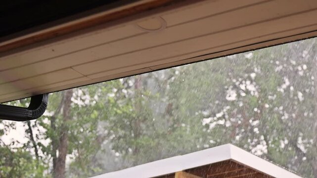 Rain pours down from eaves of building while trees sway in thunder stormy on summer day.