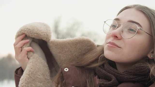 Young woman in furry trapper hat, round glasses, thick knit scarf, and padded coat adjusts winter layers in snowy urban park, then leans back with eyes closed and gentle smile in cold daylight