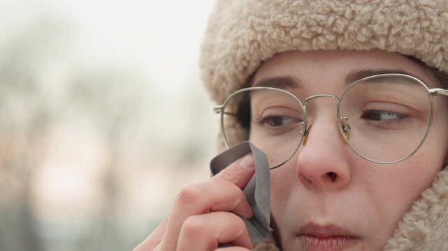 Tight portrait of young adult wearing thick fleece hat and round metal glasses, shifting hand across face, blinking, exhaling visible breath, and smiling in cold outdoor scene with soft light and