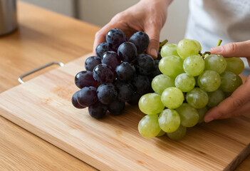 Naklejka premium Healthy eating and fresh fruit concept, hands holding green and red grapes on a wooden cutting board in a kitchen