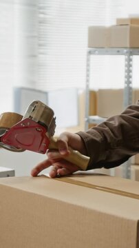 Vertical video of Caucasian young adult male sealing cardboard box with tape dispenser and sticking delivery label. Footage is ideal for e-commerce logistics and small business shipping promos