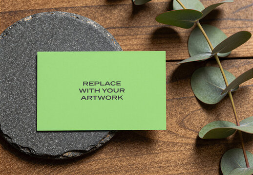 Business card mockup on a dark stone coaster with eucalyptus leaves, placed wooden surface in flat lay composition, realistic stationery branding presentation, template for graphic design