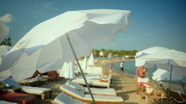 White beach parasols offering shade on a sandy beach with people relaxing and swimming in the sea on a sunny day