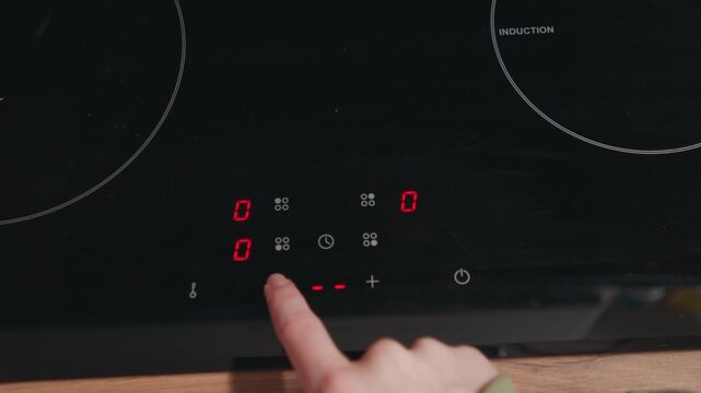 Close view of index finger pressing illuminated touch control on black glass induction cooktop, activating glowing red indicators and plus minus symbols on reflective modern kitchen surface