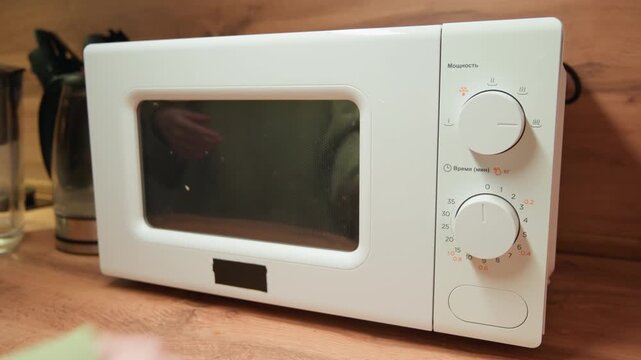 Person hand reaches to open microwave door on wooden countertop, revealing dirty interior, removed turntable, visible control knobs, and residue inside during routine kitchen cleaning and inspection