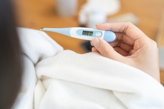 Close-up of sick Asian woman hand holding a digital thermometer showing high body temperature of 39.7 degrees celsius, female wrapping in white blanket having a severe fever at home