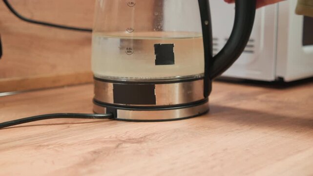 Close low-angle motion shows electric kettle descending onto round induction hotplate on wooden countertop, moving from hover to full contact, with base flush, centered alignment, and warm indoor
