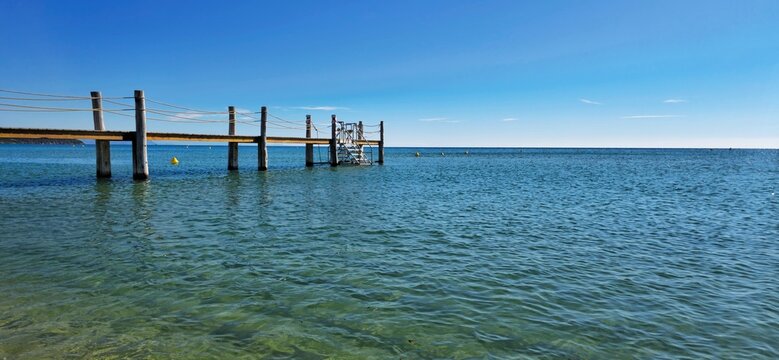 Wooden swimming pontoon with rope railings extends into the calm turquoise waters of Pampelonne Beach, Ramatuelle, near Saint-Tropez on the French Riviera, with a metal bathing ladder and coastal hill