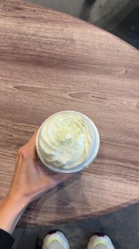 Person with a drink at the cafe table. POV video of a green matcha frappe in a takeaway cup with foam and straw. A hand moves the drink across a cafe table, pick up and check out. Copy space for text