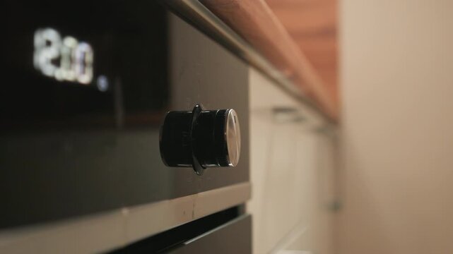 Low-angle close view of hand gripping and rotating cylindrical oven control knob beside illuminated 230 display, with soft reflections across stainless black panel and warm kitchen light