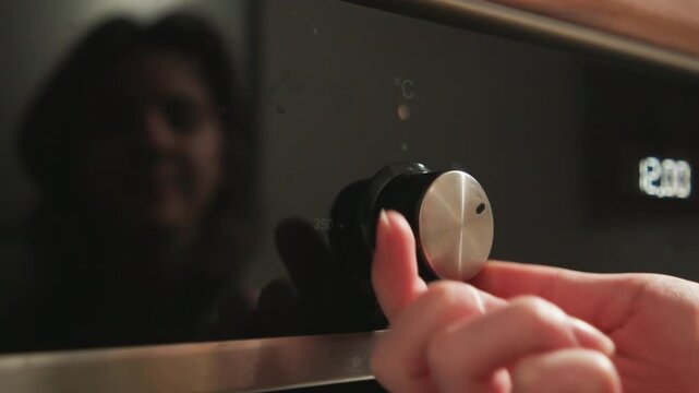 Close-up of hand turning metallic control knob on glossy black oven surface while blurred human reflection appears in panel; visible adjustment motion and fingertip position convey interaction, warm