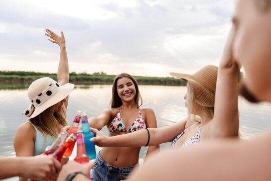 Friends Enjoying Summer Fun by the River While Holding Colorful Drinks and Celebrating Together