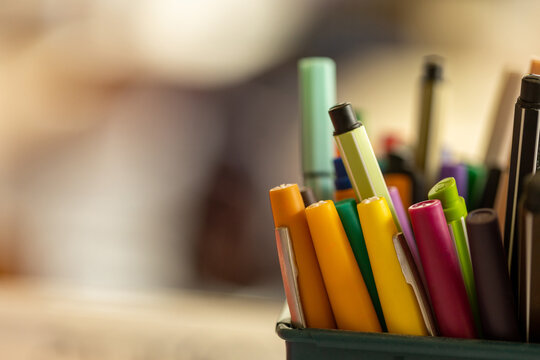 Assortiment de crayons et de fournitures scolaires color&eacute;s dans un pot, photographi&eacute; avec une faible profondeur de champ et une salle de classe floue en arri&egrave;re-plan.