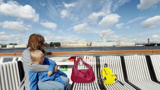 A mother lovingly hugs her young son on a boat deck in Saint Petersburg. They enjoy the scenic river view under a bright blue sky. The moment feels warm and peaceful.