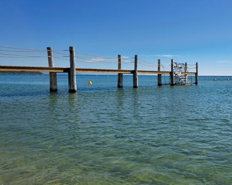 Wooden swimming pontoon with rope railings and bathing ladder stretching across the calm turquoise waters of Pampelonne Beach near Saint-Tropez, French Riviera, with coastal hills on the horizon under