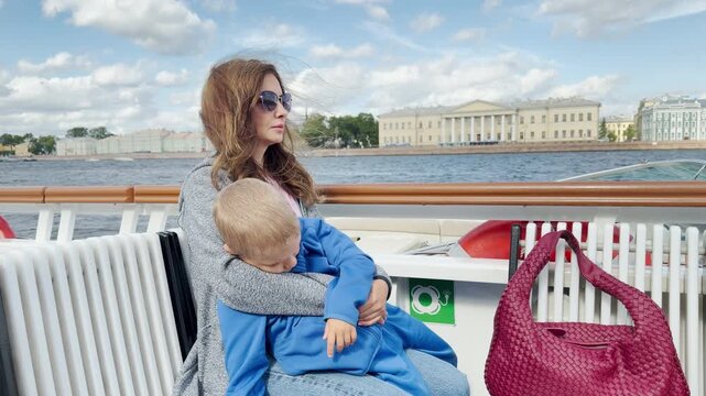 A mother holds her sleeping child resting comfortably on a boat deck in Saint Petersburg. The gentle river breeze creates a peaceful and loving atmosphere.
