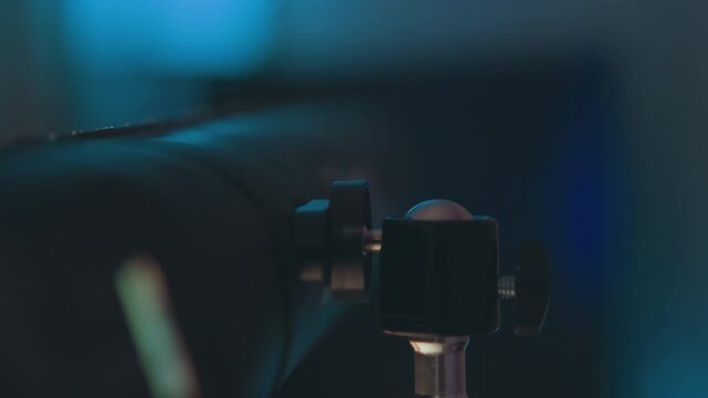 Close-up of adjustable clamp and threaded connector fixed to cylindrical boom or pole in dim studio light, with subtle tilt, shallow focus, and blurred fingertip movement hinting recent handling