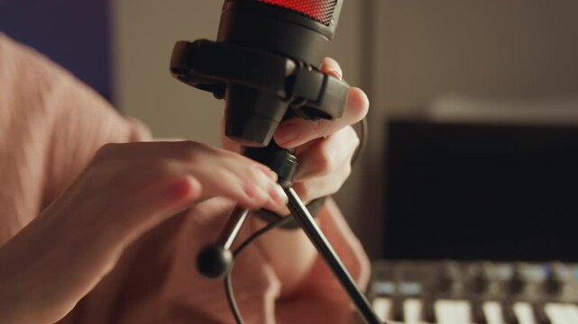 Close-up eye-level view shows hands tightening tabletop microphone in shock mount above small tripod, with cable draped across desk, warm and green mixed lighting, and careful recording setup