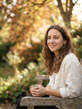 Woman sitting on a wooden bench smiling while cradling a ceramic mug outdoors