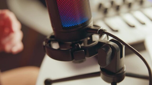 Close view of hands positioning condenser microphone inside shock mount in home studio, showing active finger movement, soft blur, glowing grille, warm light, and tidy recording workspace