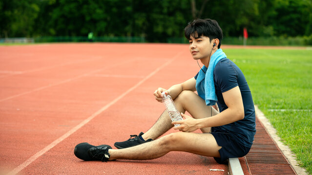 Male athlete resting on running track with water bottle, towel and earphones after training, showing hydration, recovery, fitness, exercise, endurance and outdoor sports lifestyle