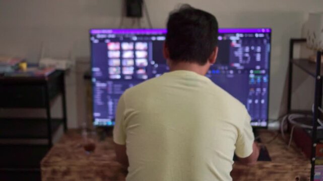 Rear view of man editing video on large computer monitor at home. Content creation, video editing workspace, lifestyle stock footage.