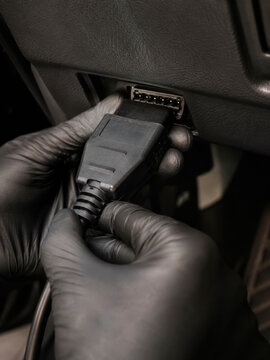 Hands wearing black gloves plug a diagnostic OBD cable into a vehicle port under the dashboard. The image emphasizes repair, inspection, and automotive diagnostics.