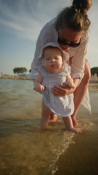Mother holding her young baby, letting baby's bare feet touch the warm ocean water on a sunny beach day