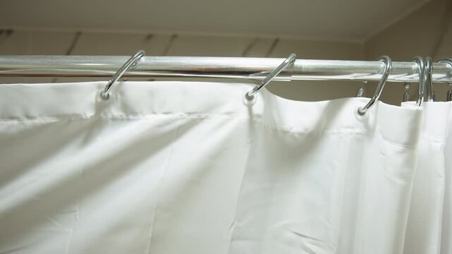Close frontal view of white shower curtain suspended from chrome rings on metal rod, with vertical folds, rust spots, and slight billowing that reveal gentle movement inside modest bathroom setting