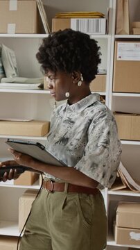 Vertical video of African American young adult woman scanning packages with barcode reader and tablet in warehouse. Clip is ideal for e-commerce logistics and small business inventory management