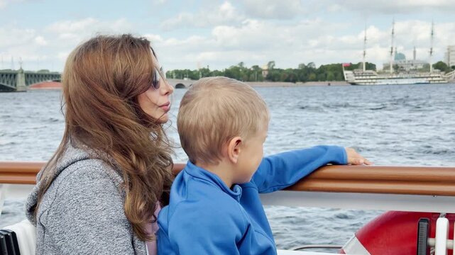 A mother and son gaze across the river from a boat deck in Saint Petersburg. They watch Trinity Bridge and distant ships under a cloudy sky. This peaceful moment fills them with wonder and calm.