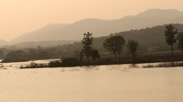 Peaceful Lake at Golden Hour with Misty Mountains and Trees Landscape. Serene River at Sunrise with Hazy Hills and Warm Sunlight . Calm Water Surface Against Hazy Mountain Silhouette During Sunset.