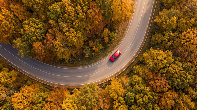 Drone view of a winding road through a vibrant autumn forest with a small red car, showcasing colorful fall foliage and a scenic seasonal landscape