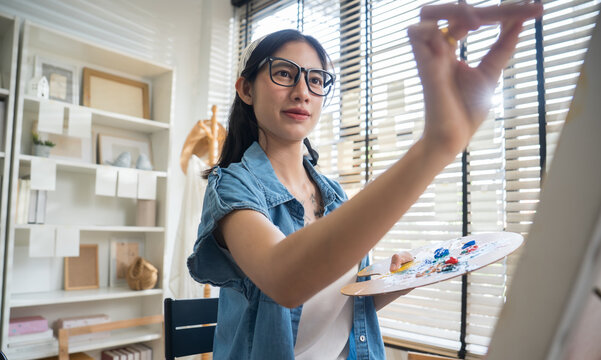 Young creative female artist sitting on a chair, painting on canvas with watercolors in a bright home art studio.