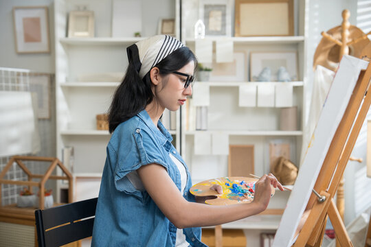 Young creative female artist sitting on a chair, painting on canvas with watercolors in a bright home art studio.